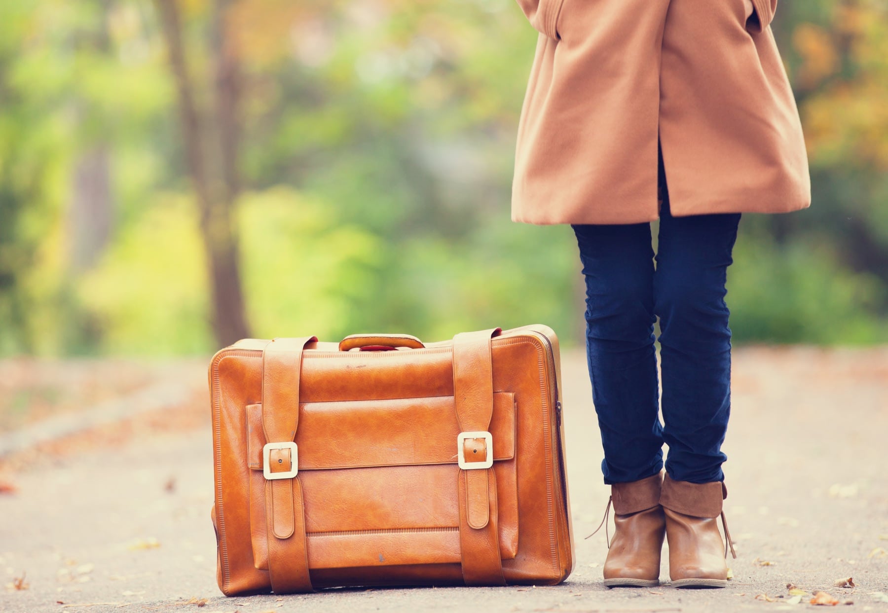 A woman wearing a beight coat and blue jeans stands next to a leather travel bag.